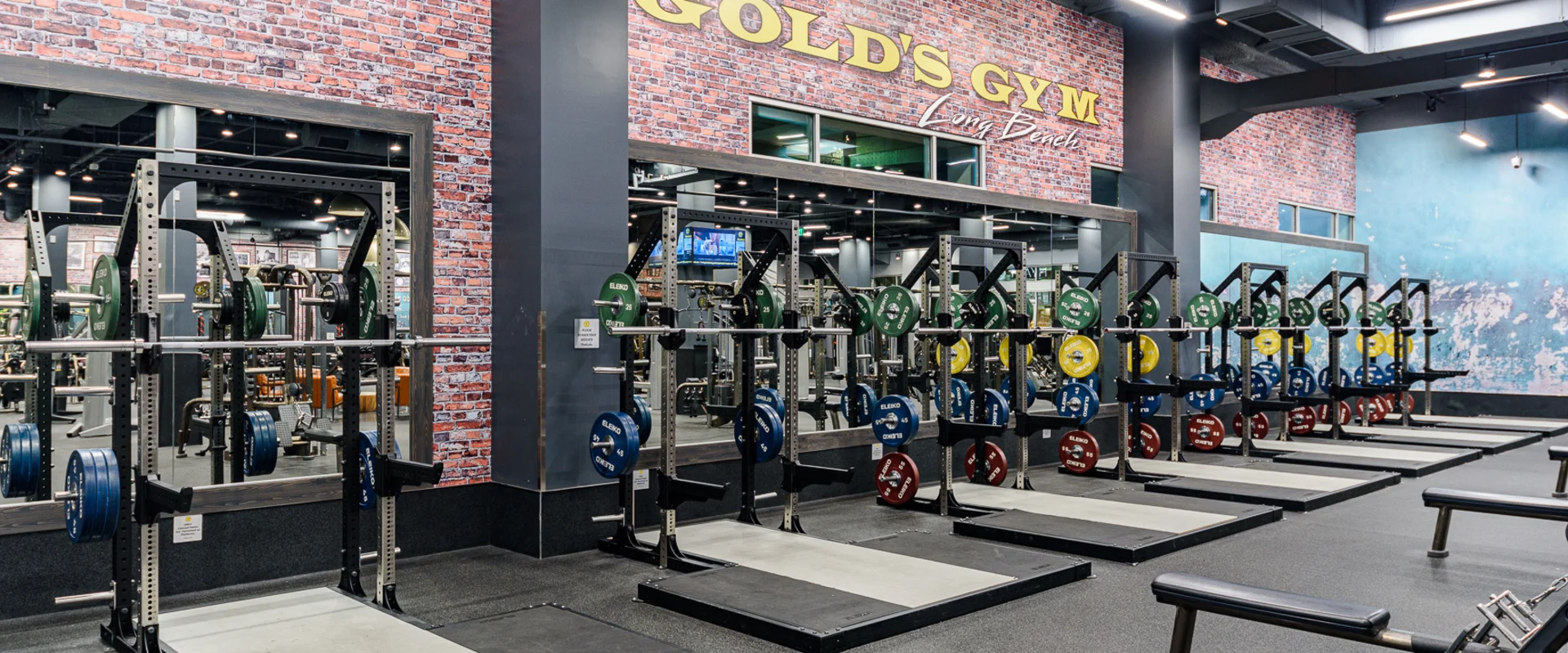Olympic lifting platforms at Gold’s Gym Long Beach, featuring multiple stations equipped with bumper plates and sturdy squat racks. Each platform is set on reinforced flooring to absorb impact, ensuring optimal support for powerlifting and Olympic-style lifts. Large mirrors line the brick-accented walls, reflecting the industrial-style gym design. Overhead LED lighting highlights the spacious setup, creating an environment built for strength and performance.
