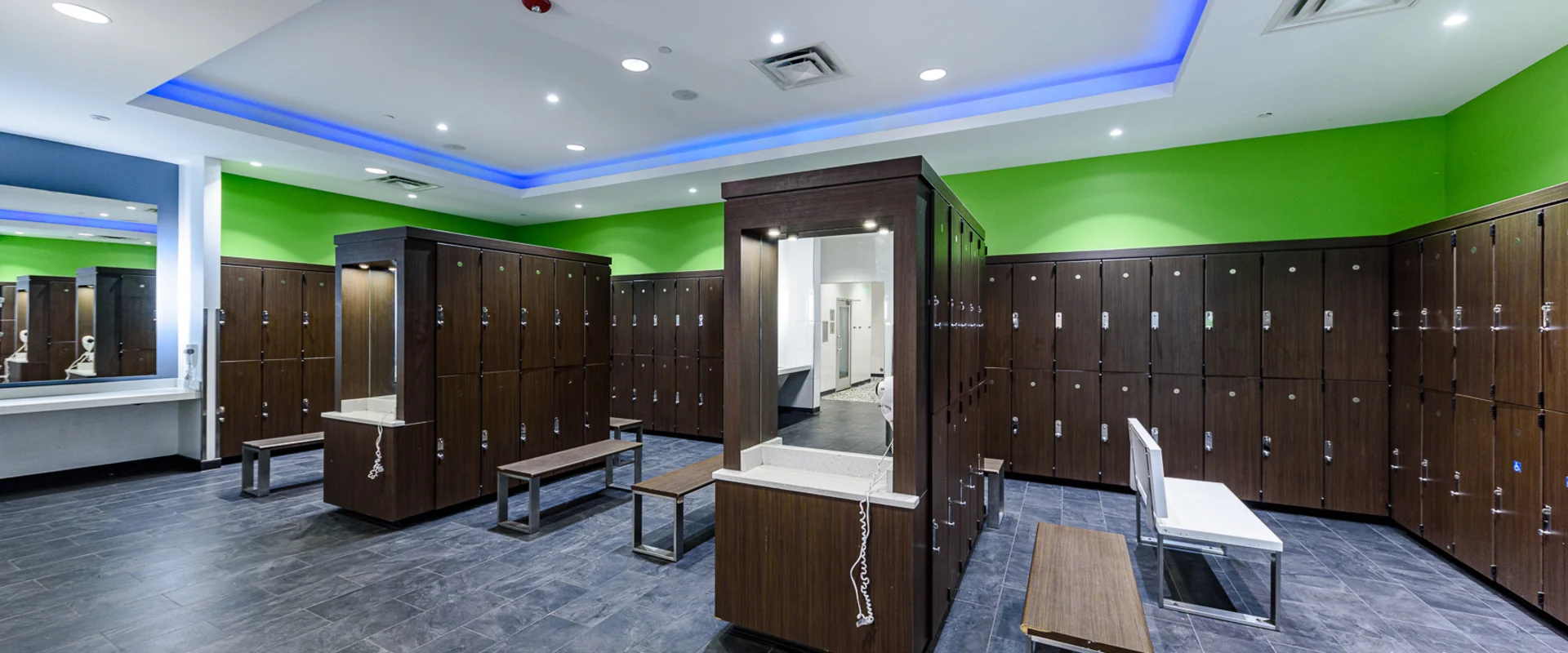 Modern locker room at Gold's Gym Garden Grove, featuring rows of dark wood lockers with silver combination locks. The space includes benches with wooden tops and metal frames, a central vanity station with a built-in mirror, and a sleek black tiled floor. The ceiling has recessed lighting with a blue LED accent strip, creating a clean and stylish look.