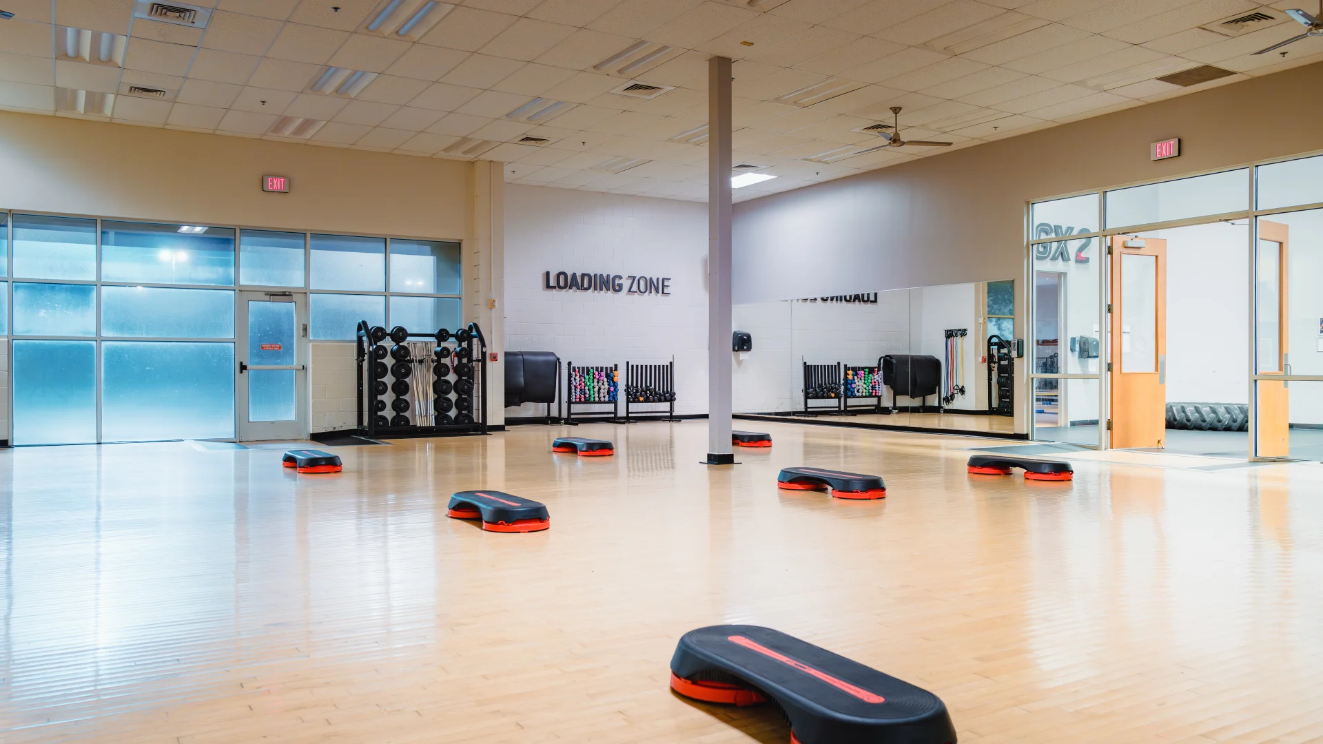 Spacious group exercise room at Gold's Gym Waco with a polished wood floor, step platforms evenly spaced, large mirrors along one wall, and storage racks holding weights and resistance bands. Frosted glass windows and double doors are visible.
