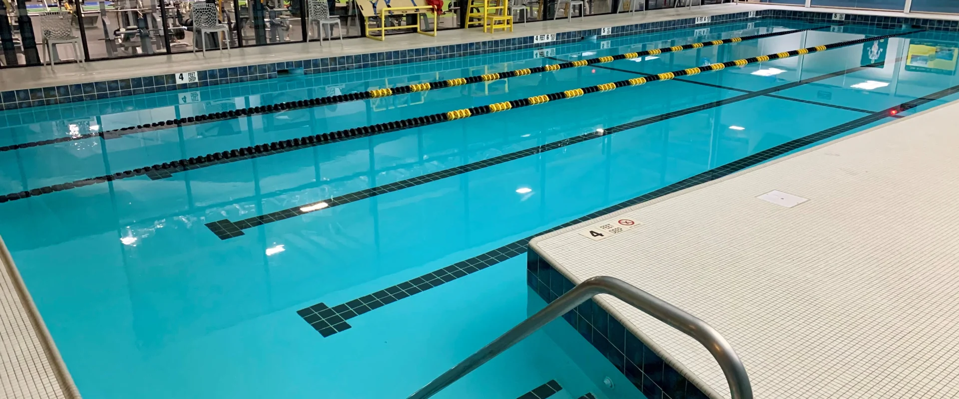 A large indoor swimming pool with clear blue water, surrounded by tiled walls and a gym area visible in the background.