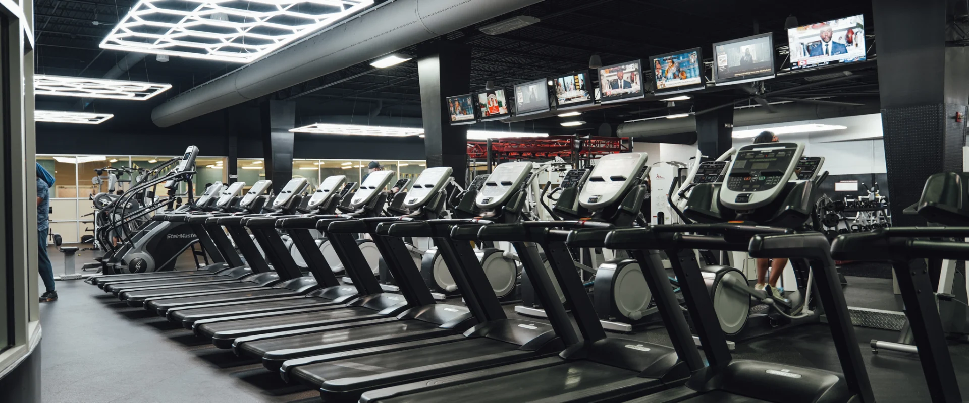 A row of high-end treadmills and elliptical machines at Gold's Gym Brassfield, positioned under sleek industrial lighting. Multiple overhead TVs display news and sports channels, creating an engaging workout environment. The modern black-and-gray gym aesthetic is complemented by exposed ductwork and geometric LED ceiling lights.