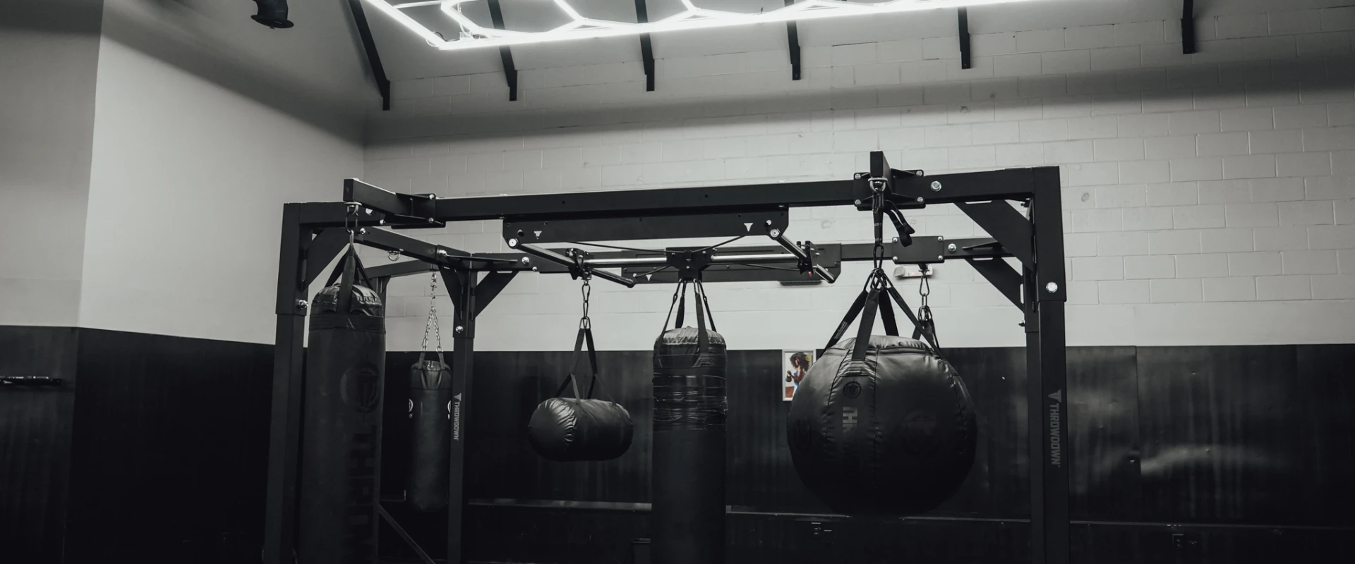 A boxing training area at Gold’s Gym Randleman featuring a steel rig with hanging heavy bags and training bags. Overhead LED grid lighting illuminates the open floor space, with dark wall finishes and an industrial training environment.