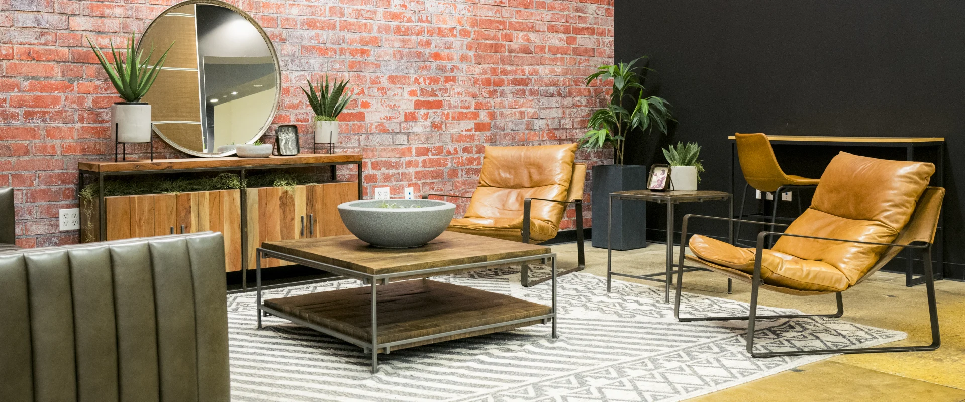 Lounge area at Gold's Gym Waxahachie featuring leather chairs, a wooden coffee table, and a matching credenza with a round mirror mounted on a red brick accent wall. The space includes potted plants, a patterned area rug, and a desk with additional seating, creating a stylish and comfortable environment for members.