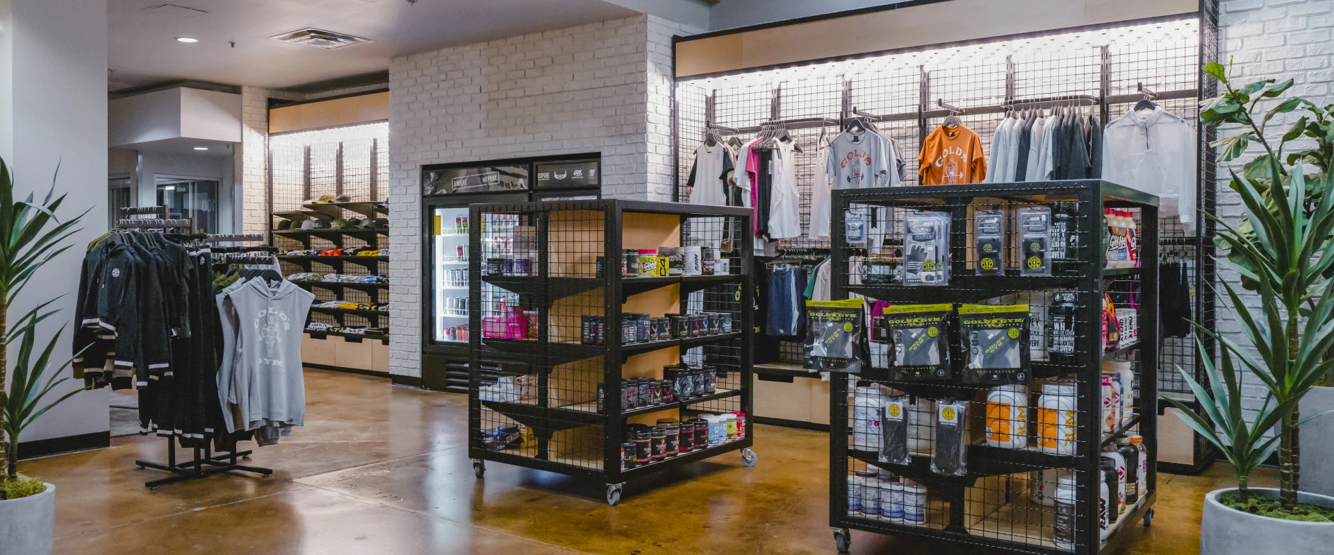 A gym pro shop featuring racks of clothing, shelves with fitness supplements and accessories, and a fridge with drinks, set in a modern, well-lit space.