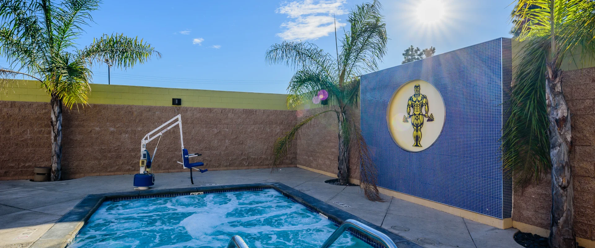 Outdoor hot tub area at Gold's Gym Garden Grove, featuring bubbling water, stainless steel handrails, and an ADA-compliant chair lift. The space is enclosed by a brick privacy wall, with tropical palm trees and a large tiled logo of Gold's Gym. The sun casts a bright glow over the serene setting.