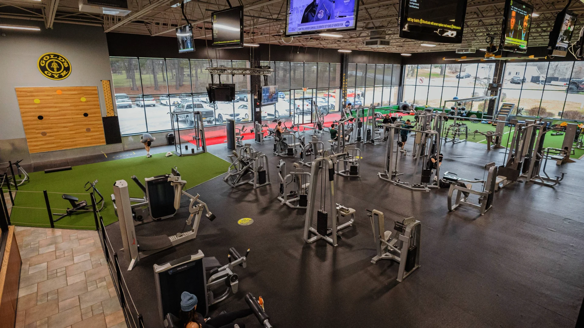 Gold's Gym New York (Poughkeepsie) gym floor showcasing a spacious layout with resistance machines, functional training turf, and a dedicated stretching area. Large windows flood the space with natural light, while multiple flat-screen TVs hang from the ceiling. The turf section features sleds, kettlebells, and functional equipment, with a climbing wall accenting the back wall near the Gold’s Gym logo.