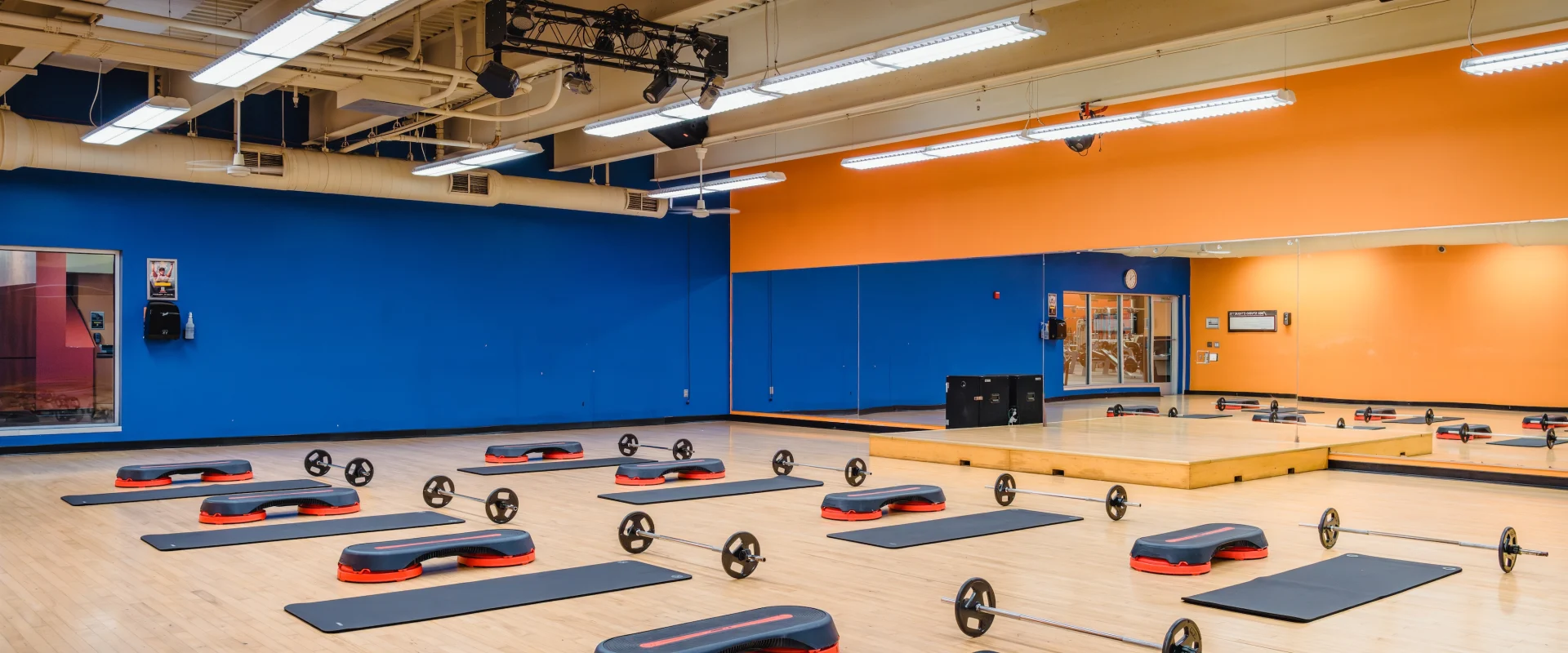 The group exercise room at Gold's Gym Fiesta Trails with orange and blue walls, mirrored panel, and hardwood floors. Equipment includes mats, barbells, and step platforms neatly arranged.
