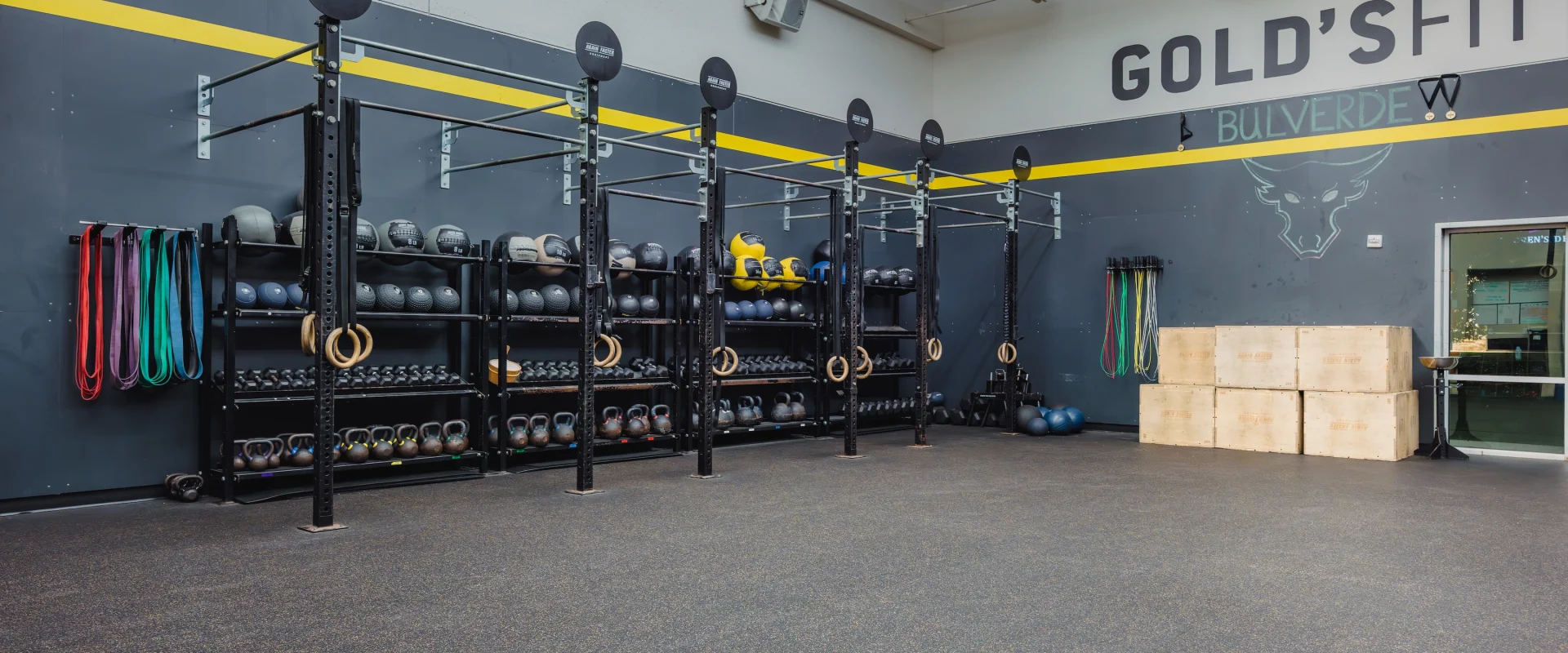 Spacious gym interior with wall-mounted racks holding kettlebells, medicine balls, and resistance bands. Bright, organized, and motivating atmosphere.