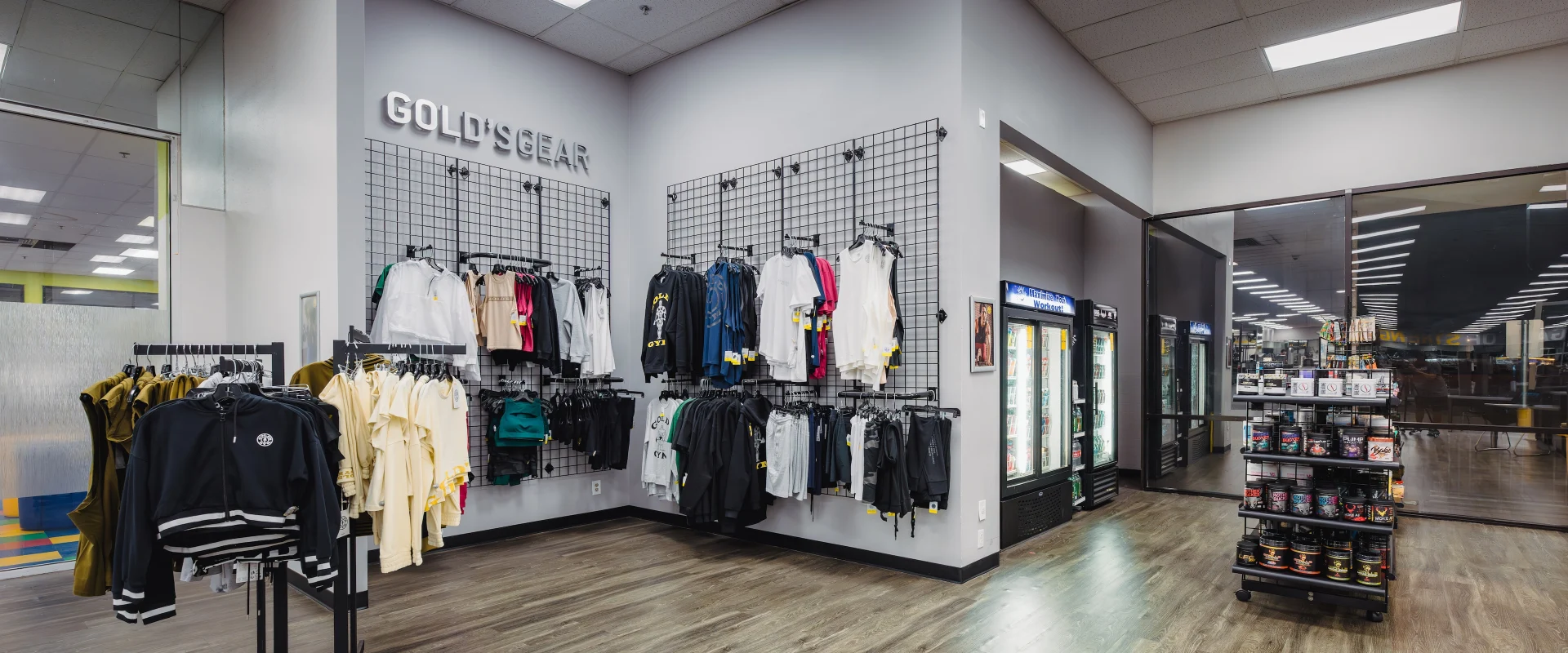 Pro shop at Gold’s Gym in Austin (South Central), featuring Gold’s Gear apparel displayed on wall-mounted racks and freestanding hangers with hoodies, t-shirts, and workout gear. The space includes refrigerated coolers stocked with beverages and a display shelf featuring supplements and protein products. Wood-style flooring and bright overhead lighting create an inviting retail area within the gym.