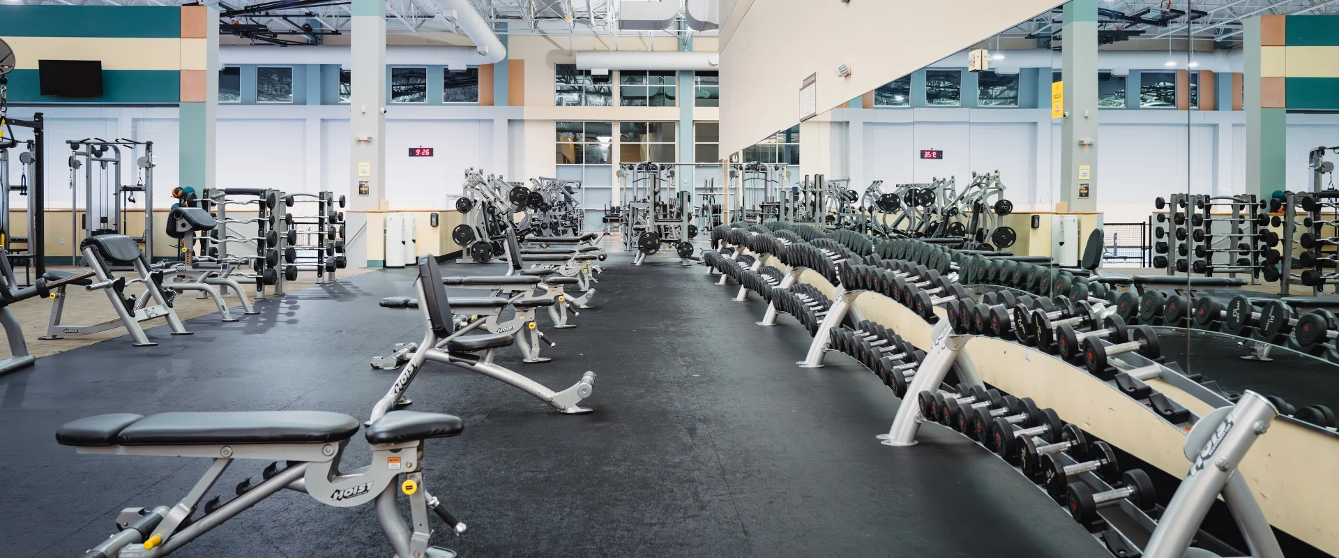 Spacious gym with rows of dumbbells and weight benches on a black floor. Bright lighting, tall mirrors, and high ceilings create an open atmosphere.