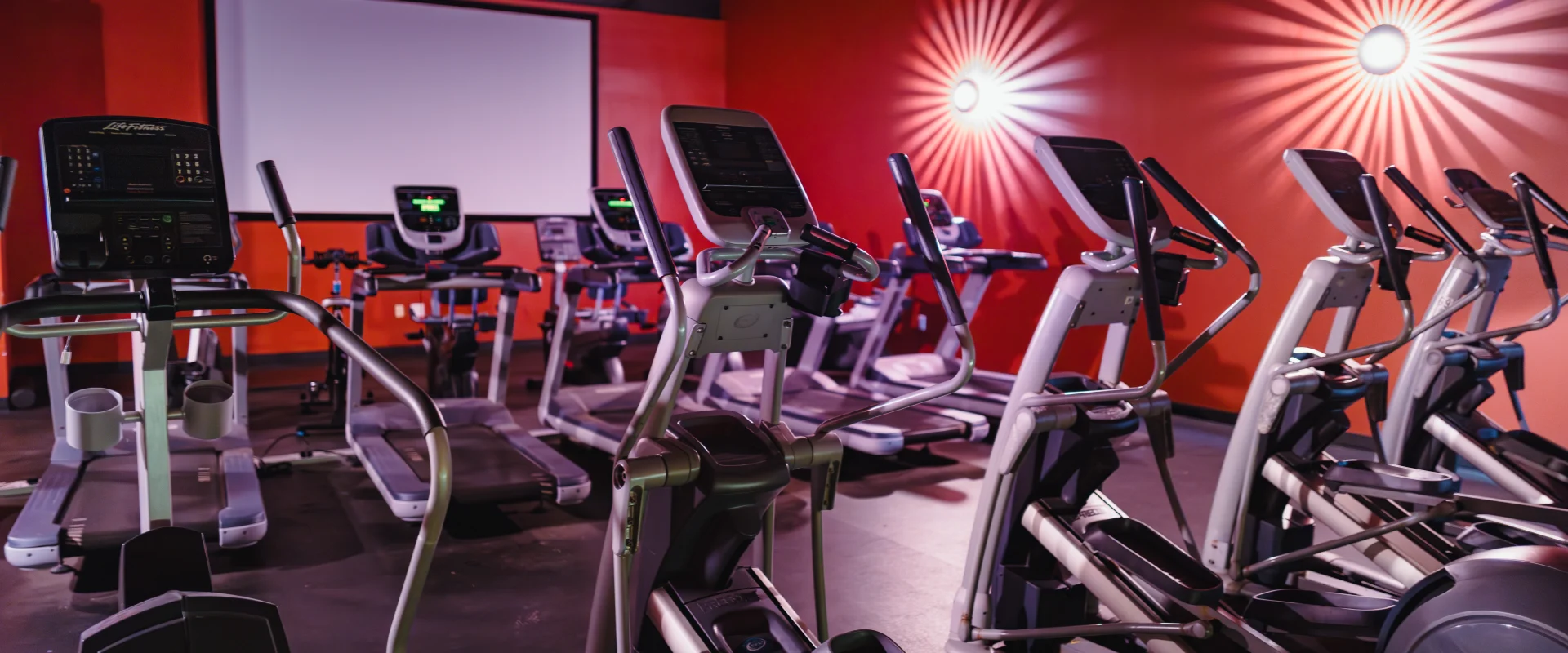 Cardio Cinema at Gold's Gym Austin (Bee Caves) featuring rows of cardio equipment in a theater-style setup. The room includes treadmills and elliptical machines facing a large projection screen, with a bold red wall and decorative wall sconces creating a cinematic atmosphere.