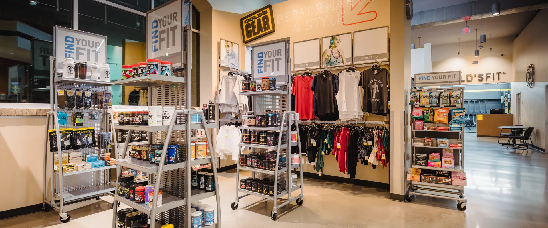 Pro Shop at Gold's Gym Austin (Downtown) showcasing shelves stocked with workout supplements, protein powders, energy bars, and fitness accessories. The display also includes racks of gym apparel such as hoodies, tank tops, and leggings. Bright lighting highlights the clean, modern retail space with promotional signs and a polished concrete floor.