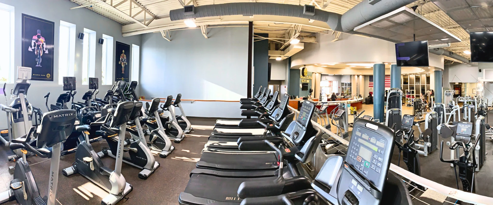 Cardio deck in a Smithtown gym featuring rows of treadmills, stationary bikes, and ellipticals arranged under bright natural light from tall windows. The space includes motivational fitness posters on the walls, with a view of the gym’s reception desk and strength training area in the background. High ceilings and an open layout enhance the modern, spacious atmosphere.