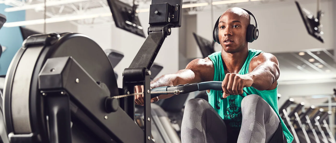 A person wearing a green tank top and headphones is seated on a fitness machine in a gym setting, with various exercise equipment visible in the background.