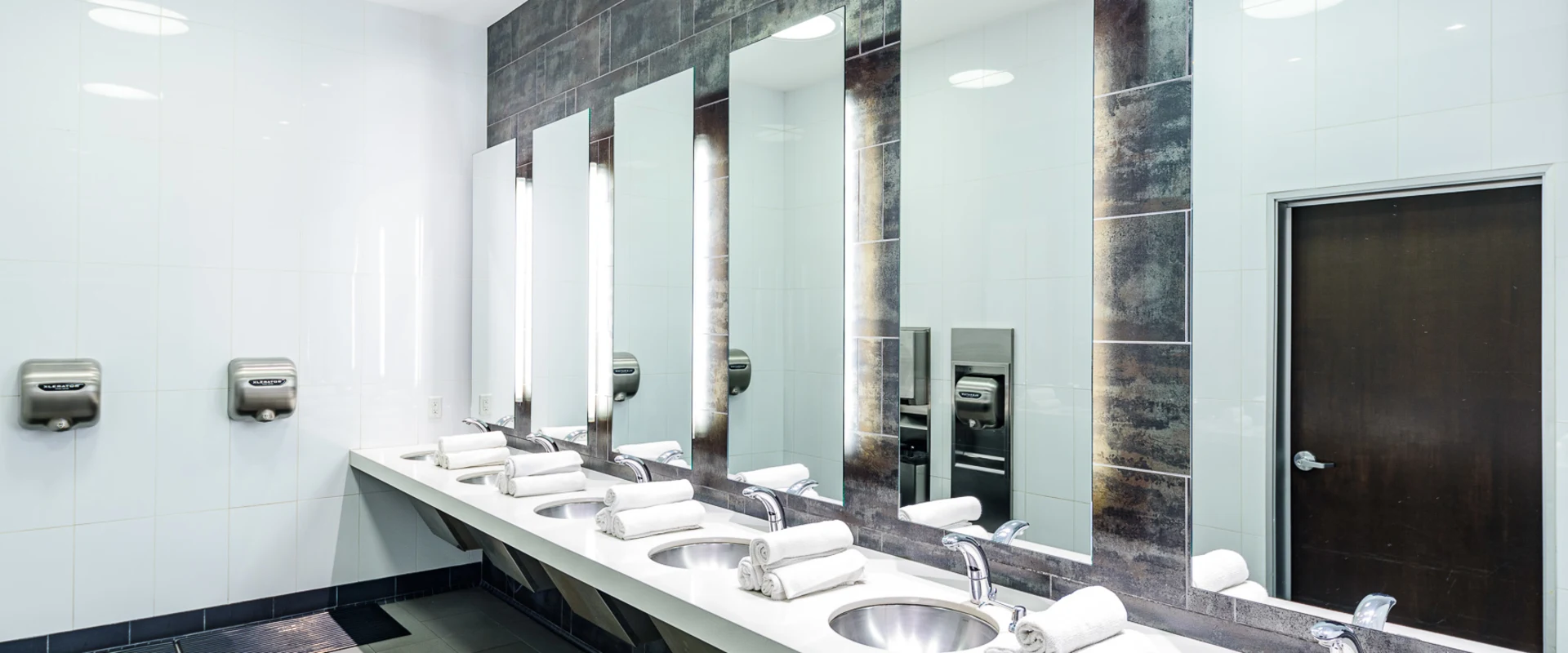 Sink area at Gold’s Gym Hollywood, featuring a long white countertop with multiple stainless steel sinks and modern chrome faucets. Stacks of neatly rolled white towels are placed beside each sink. Large mirrors with integrated vertical lighting extend across the wall, framed by dark textured tiles. The surrounding walls are covered in glossy white tiles. Stainless steel hand dryers are mounted on the wall. The floor consists of large square tiles with a central drain. A dark wooden door is visible on the right side.
