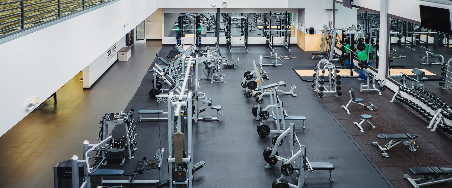 Wide-angle view of the free weights area at Gold's Gym Oklahoma City (Moore), featuring benches, barbells, and weightlifting machines on a spacious gym floor with additional cardio equipment visible on the upper level.