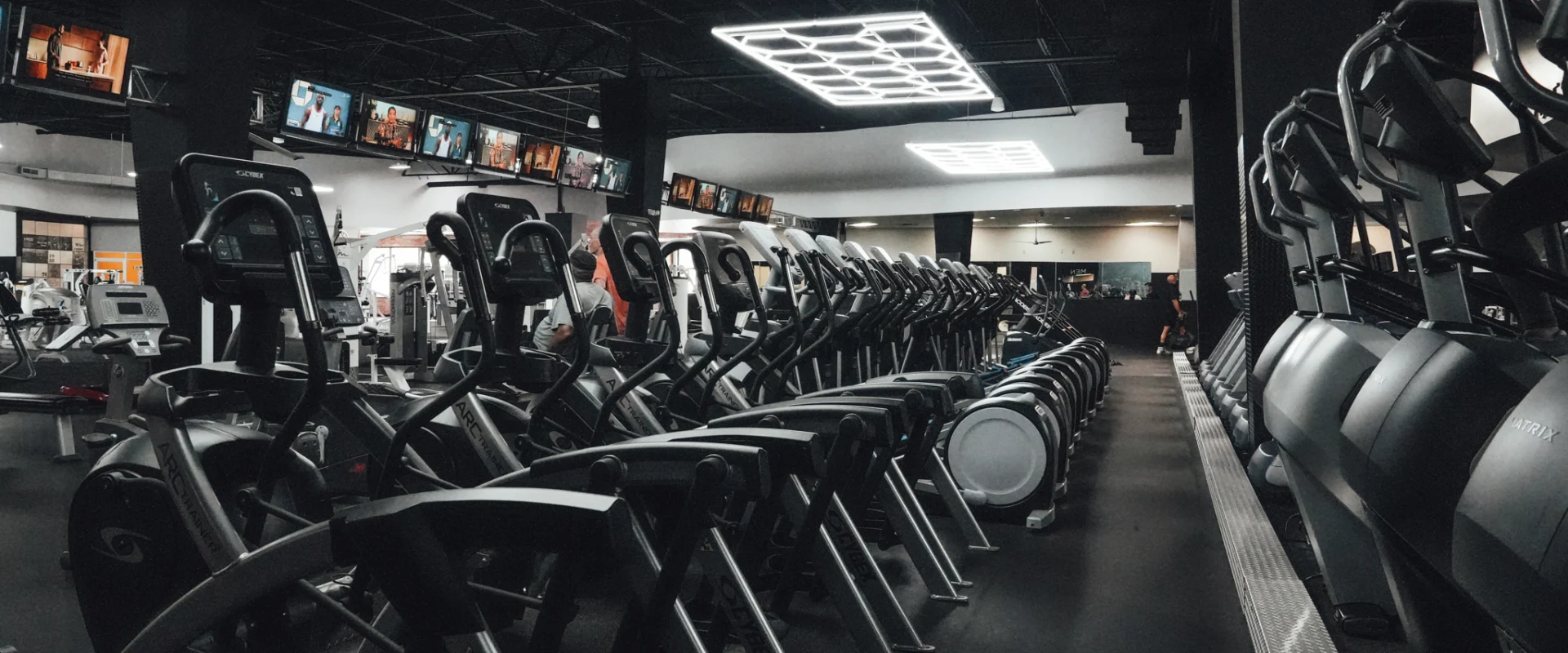 A long row of elliptical cardio machines at Gold’s Gym Randleman positioned beneath geometric LED ceiling lighting, with televisions mounted overhead across the cardio zone.