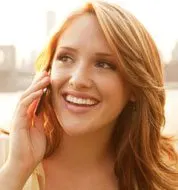 A smiling woman with long, wavy blonde hair is talking on a mobile phone against a blurred urban background.