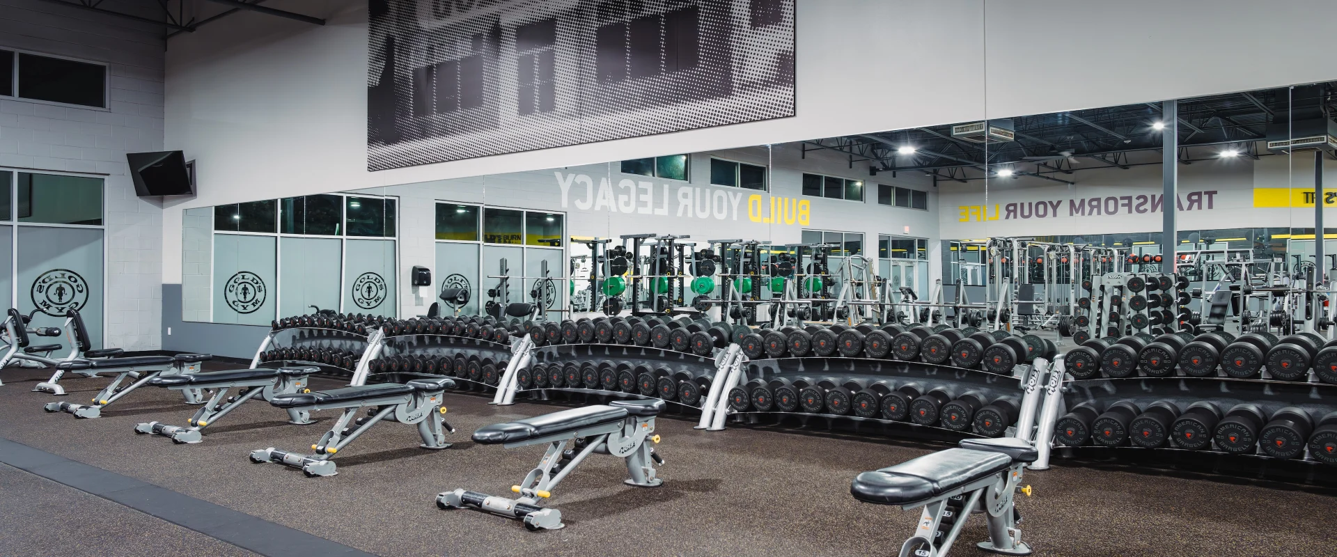The dumbbell area at Gold's Gym in Texas (Slaughter & South Congress) includes multiple racks of dumbbells neatly organized by weight. Adjustable benches are positioned for strength training, with large wall mirrors providing visibility for form checks. Motivational signage adds energy to the space.
