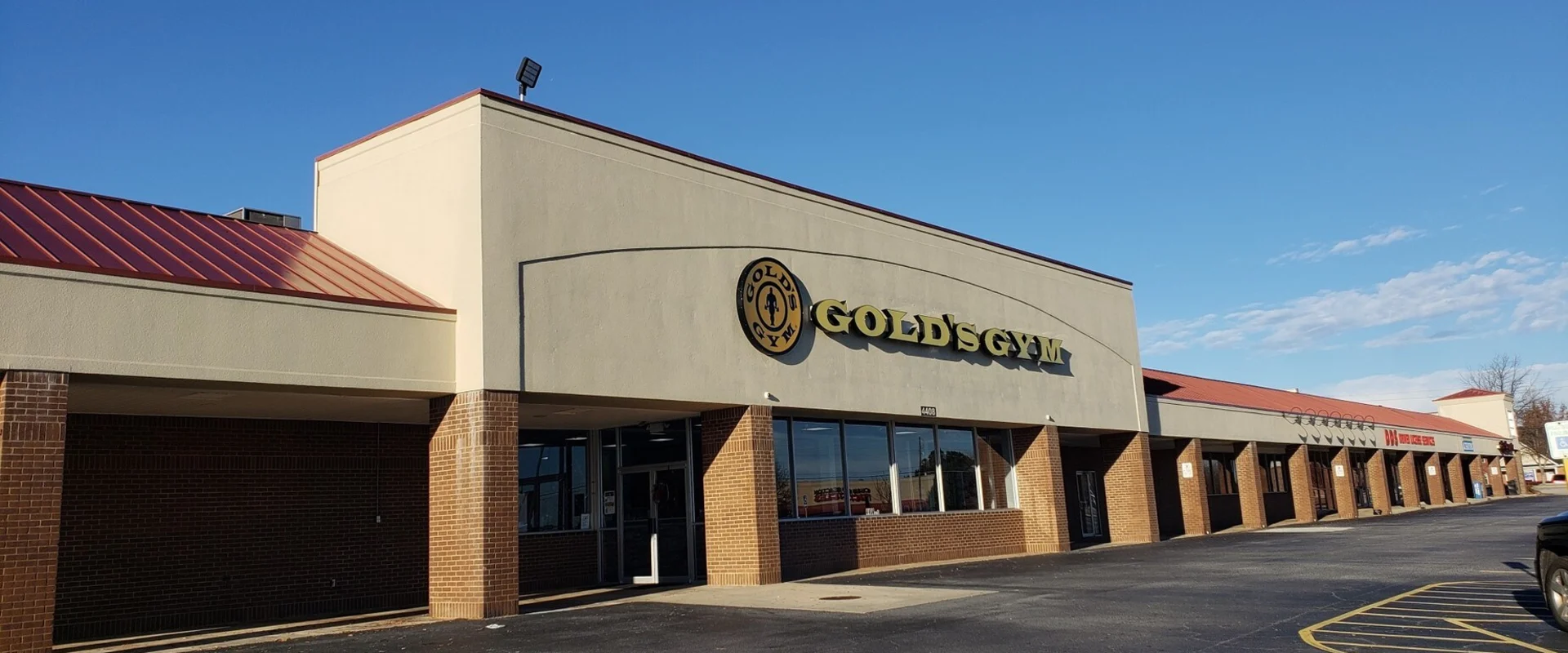 Exterior of Gold’s Gym Evans with yellow signage on tan and brick building under a bright blue sky.