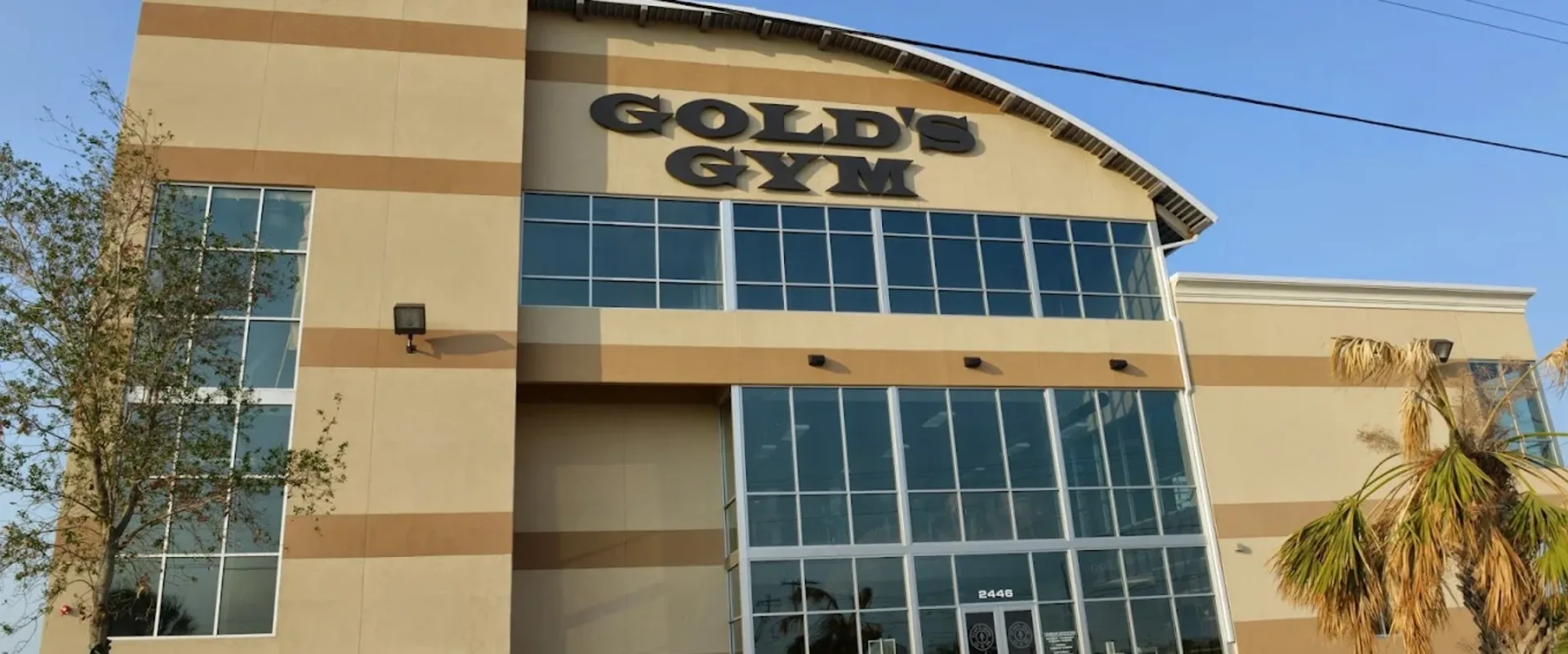 Exterior of Gold’s Gym Laredo North Central with tall windows and tan facade under blue sky.