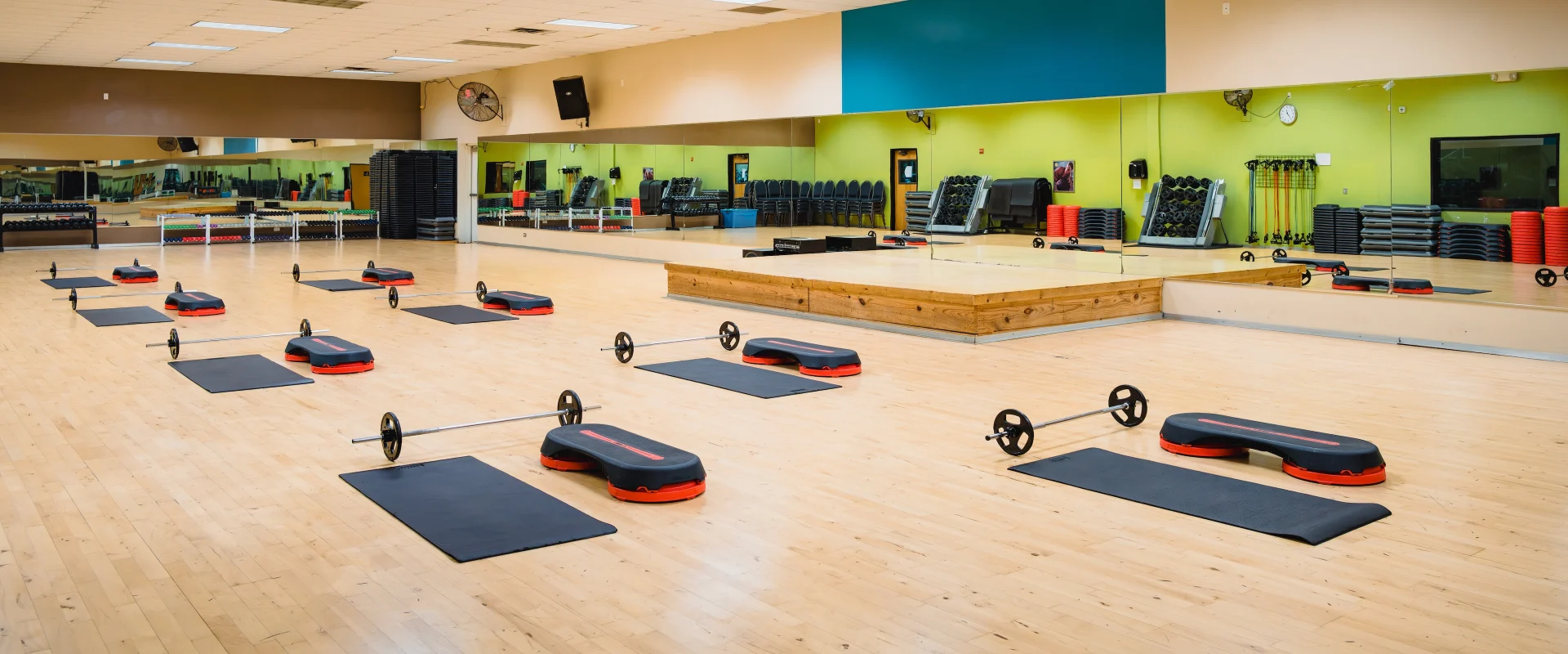A spacious group exercise studio at Gold’s Gym in Austin (South) features a light wood floor and mirrored walls reflecting the open layout. Black mats with red and black aerobic steps and barbell sets are evenly spaced across the floor, ready for a class. A wooden platform stage sits at the front for instructors, with a variety of fitness equipment neatly stacked along the walls, including resistance bands, weight plates, and additional steps. Bright overhead lighting and vibrant green and blue accent walls create an energetic and inviting atmosphere.