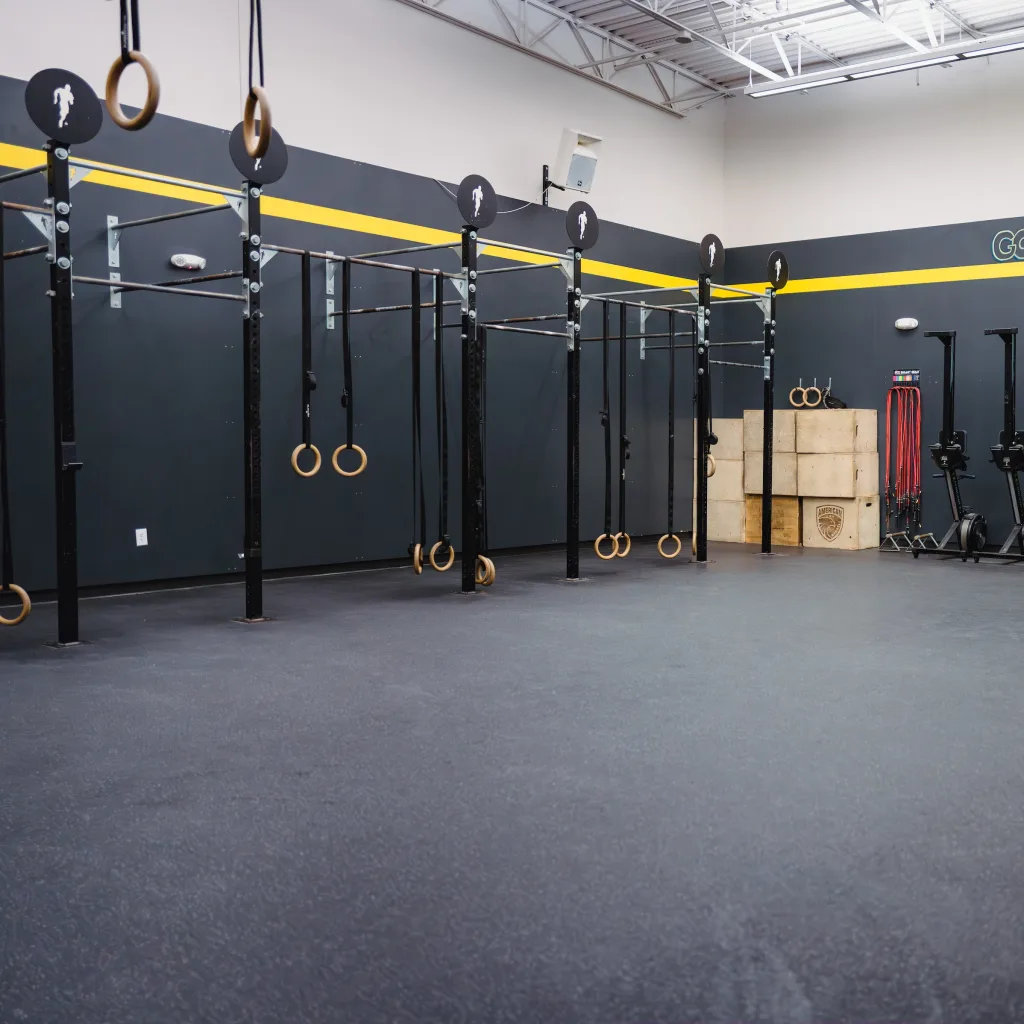 Spacious gym with black-and-yellow walls, pull-up bars, gymnastic rings, stacked boxes, and row machines lined up, creating a focused workout vibe.