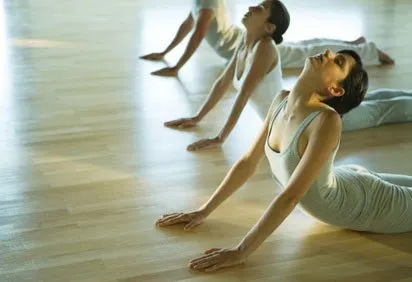 The image depicts a group of people, likely dancers, performing a synchronized movement on a wooden floor with a bright, diffused light in the background.