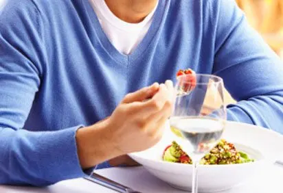 A person in a blue sweater is holding a glass of white wine and a bowl of what appears to be a salad or other healthy dish.