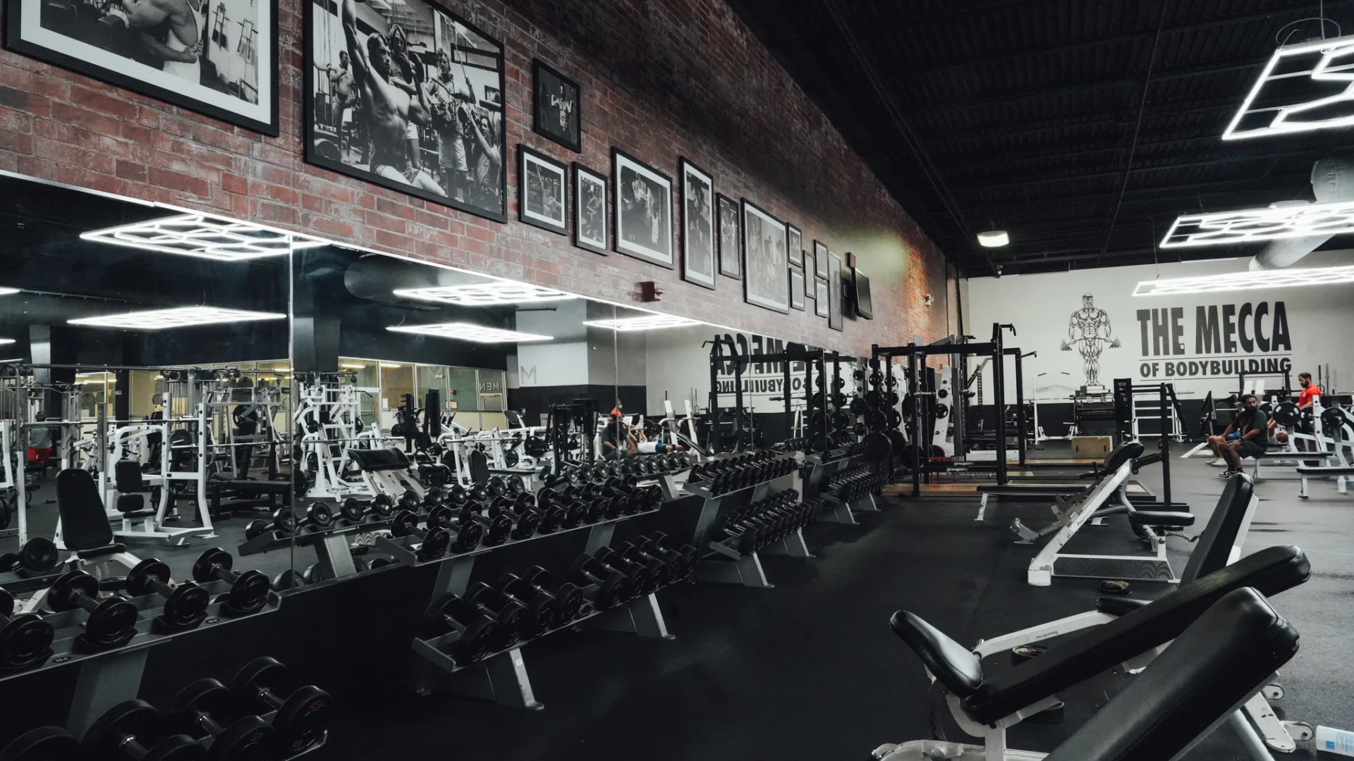 A spacious free-weight section at Gold's Gym Brassfield, lined with dumbbells on angled racks. Benches and squat racks are available for strength training. A large mirror spans the wall, reflecting the industrial-style red brick accent wall decorated with black-and-white bodybuilding photos.