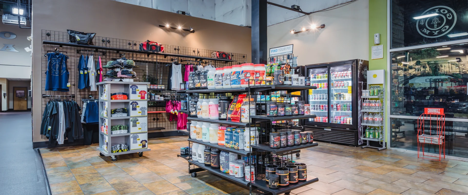 The pro shop at Gold’s Gym in Austin (South) features neatly organized shelves stocked with various supplements, protein powders, and energy drinks. A central display showcases large containers of workout supplements, while a refrigerated cooler in the background holds bottled beverages. To the left, racks display gym apparel, including tank tops, t-shirts, and hoodies, with gym bags and accessories arranged above. The space has tiled flooring and large windows allowing natural light to brighten the area. A small merchandise stand with colorful t-shirts is positioned near the center.