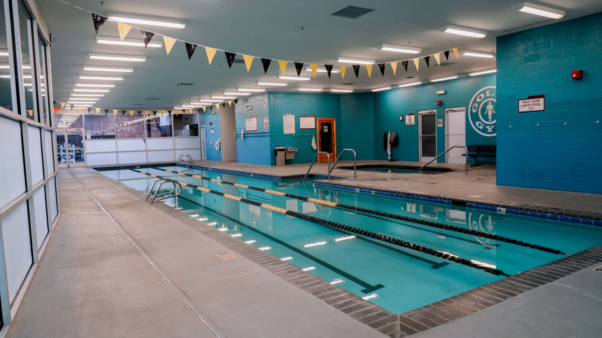 An indoor lap pool at Gold's Gym Brassfield, surrounded by bright turquoise walls and a clean tile deck. Black and gold pennant flags hang from the ceiling, while large windows let in natural light. The pool is equipped with handrails and floating lane dividers for structured swim training.