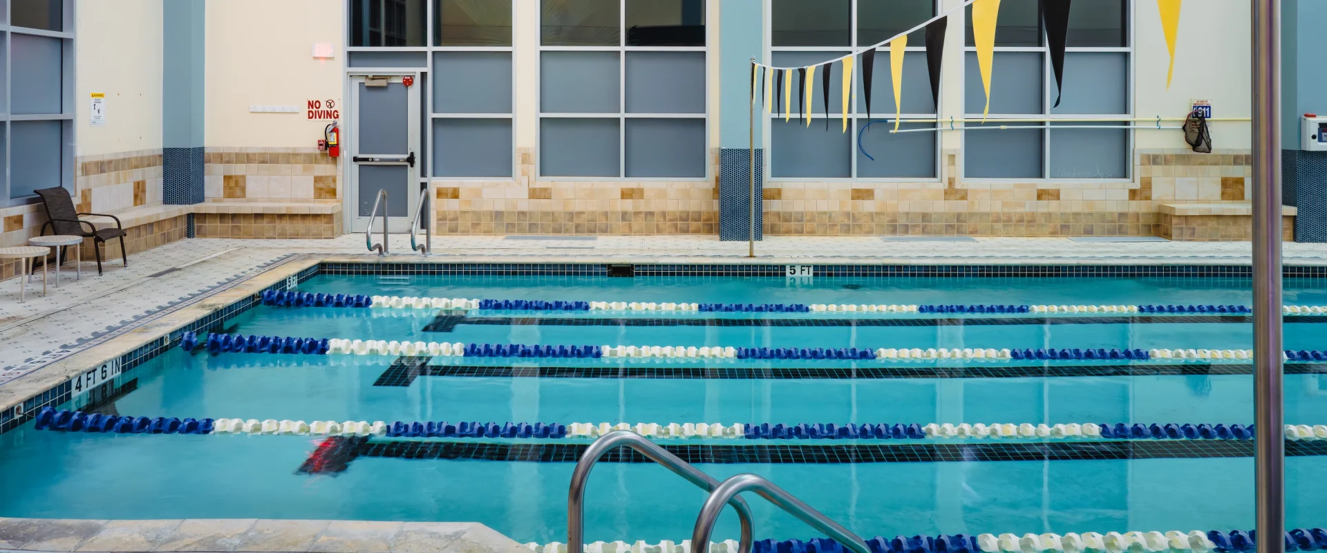 Indoor swimming pool with clear blue water, divided by blue and white lane markers. Yellow and black pennants hang above, with a serene and inviting atmosphere.