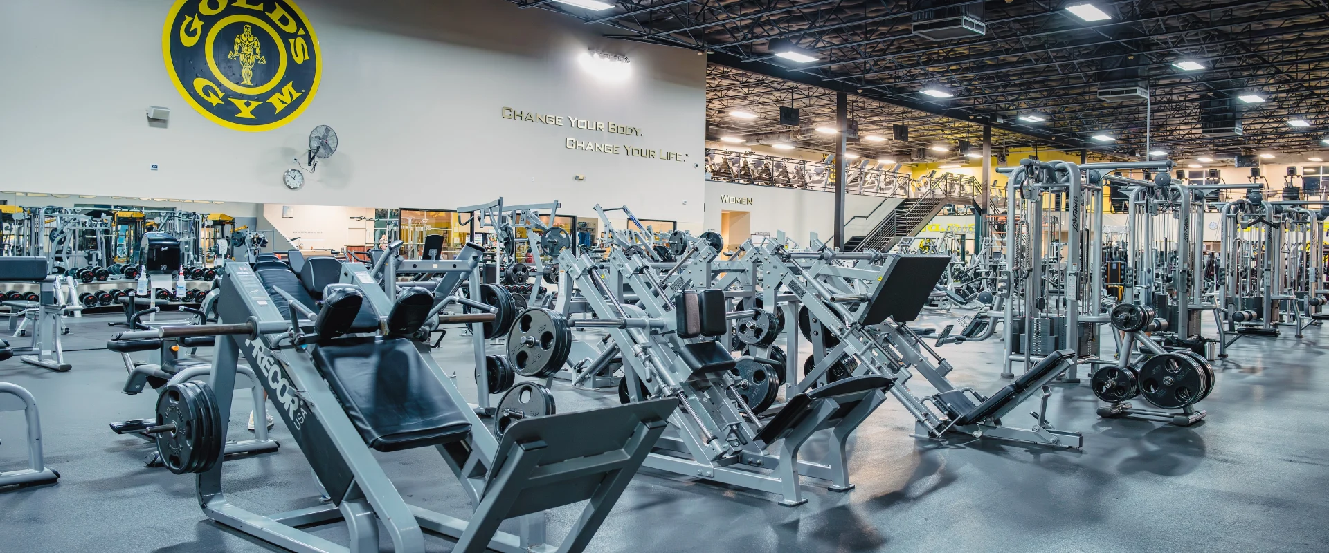 The free weights area at Gold’s Gym in Austin (South) features an array of plate-loaded machines and weight benches neatly arranged across the spacious floor. Heavy-duty leg press and hack squat machines dominate the foreground, loaded with black weight plates. Behind them, rows of cable machines, benches, and additional free weight equipment stretch across the gym. A large Gold’s Gym logo is displayed prominently on the wall above mirrored panels that reflect the extensive equipment. Overhead, bright lighting and exposed industrial ceilings give the space an open, modern feel.
