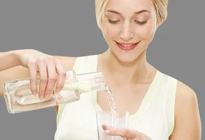 A smiling woman in a white top is pouring water from a glass container into her hands.
