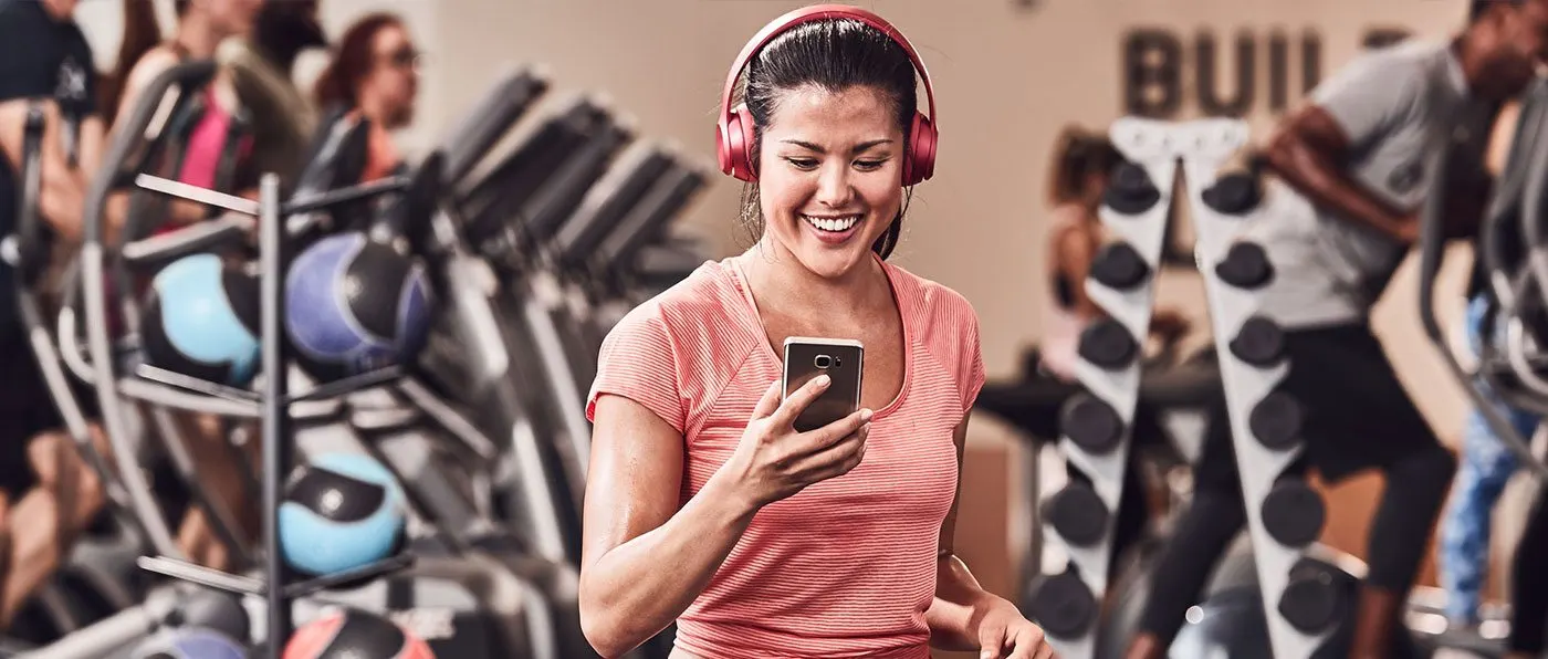 A young woman wearing pink headphones and a pink shirt is smiling while holding a smartphone in front of a colorful graffiti-covered wall.