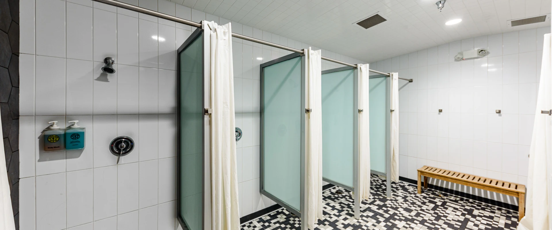 A clean and modern gym shower area with individual stalls separated by frosted glass panels and white privacy curtains. The walls are lined with glossy white tiles, while the black-and-white mosaic tile floor adds a stylish contrast. Soap dispensers and a wooden bench are provided for convenience.