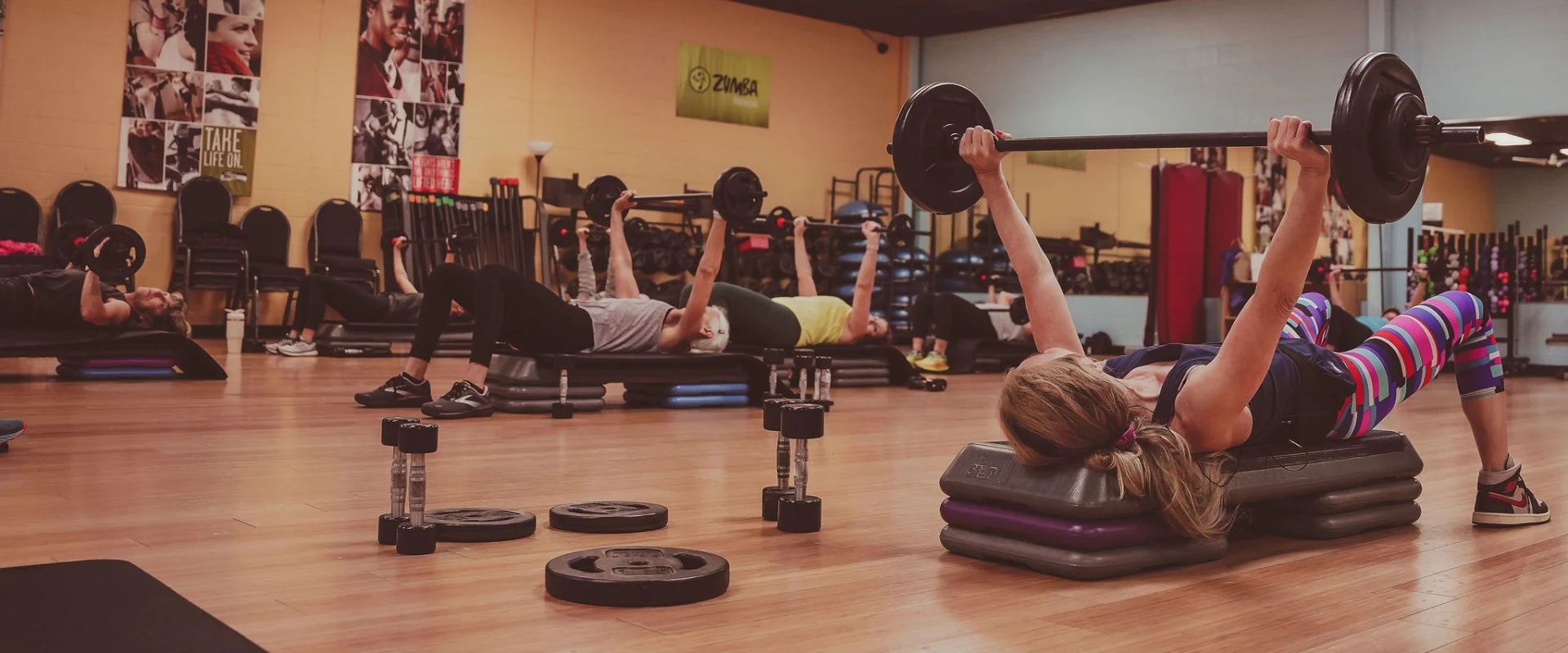 Gold's Gym Jackson, Tennessee, group exercise class with participants performing a barbell chest press on aerobic steps. The room features wooden flooring, mirrors, and fitness equipment, with motivational banners on the walls.