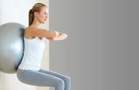 A young woman in a white tank top and gray leggings stands against a plain gray wall, her arms crossed as she gazes off to the side.