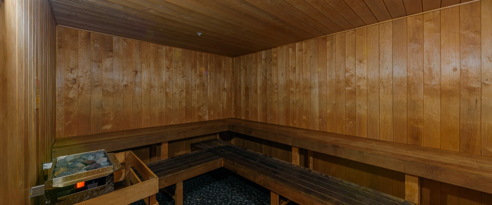 The sauna at Gold’s Gym Cerritos features wooden walls, benches, and a ceiling, creating a warm and relaxing atmosphere. A heating unit filled with sauna stones sits in the corner, and the floor is covered in black mosaic tiles. A single wall-mounted light provides soft illumination.