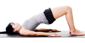 The image shows a woman in a yoga pose, lying on her back with her legs raised and her body arched. The background is plain and white, allowing the focus to be on the woman's form and the yoga pose.