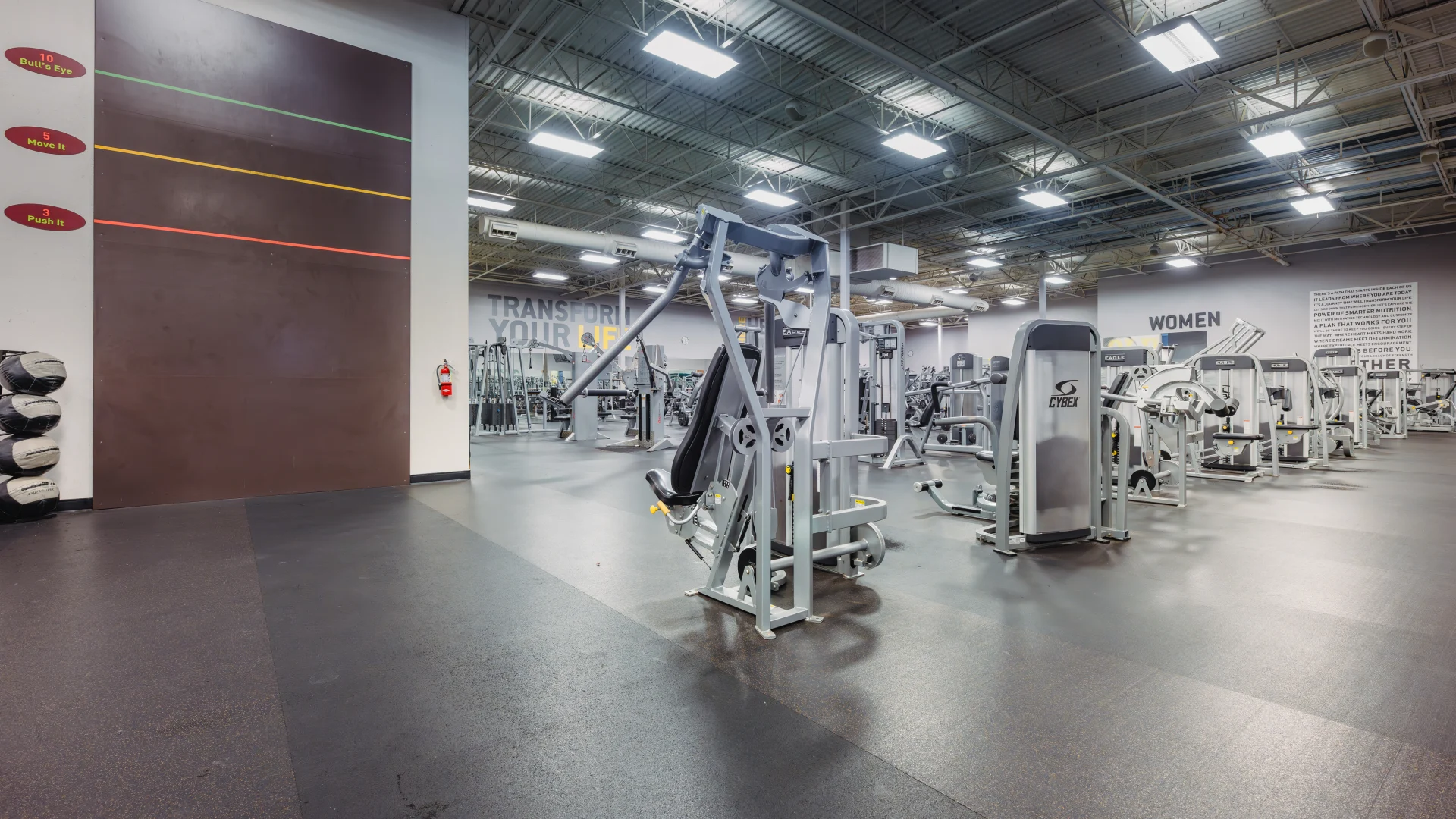 Machine weight area at Gold's Gym Waco featuring rows of strength-training machines on black rubber flooring. A tall brown plyometric wall with colored target zones stands on the left, with medicine balls stacked nearby. Overhead lighting and motivational wall text create a bright, focused workout environment.