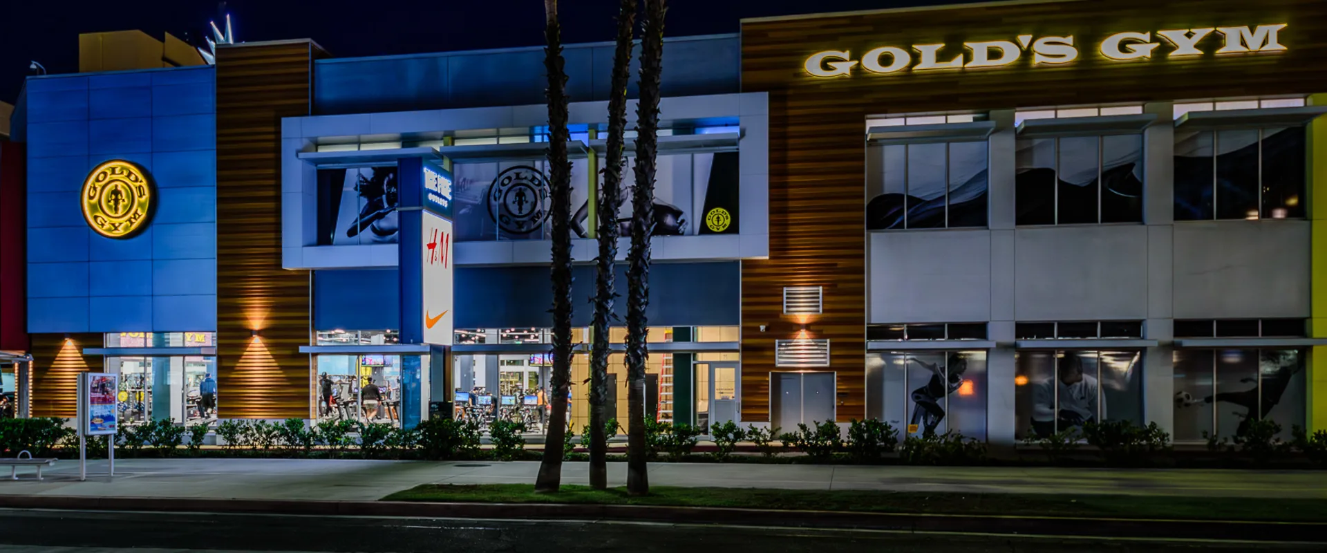 Exterior of Gold’s Gym Long Beach at night, illuminated by bright signage and warm interior lighting. The modern facade features a mix of wood paneling and sleek metal accents, with large windows revealing the gym's active interior. The entrance is flanked by palm trees, and nearby retail stores contribute to the vibrant urban atmosphere. The gym's branding stands out against the dark sky, creating a striking nighttime presence.