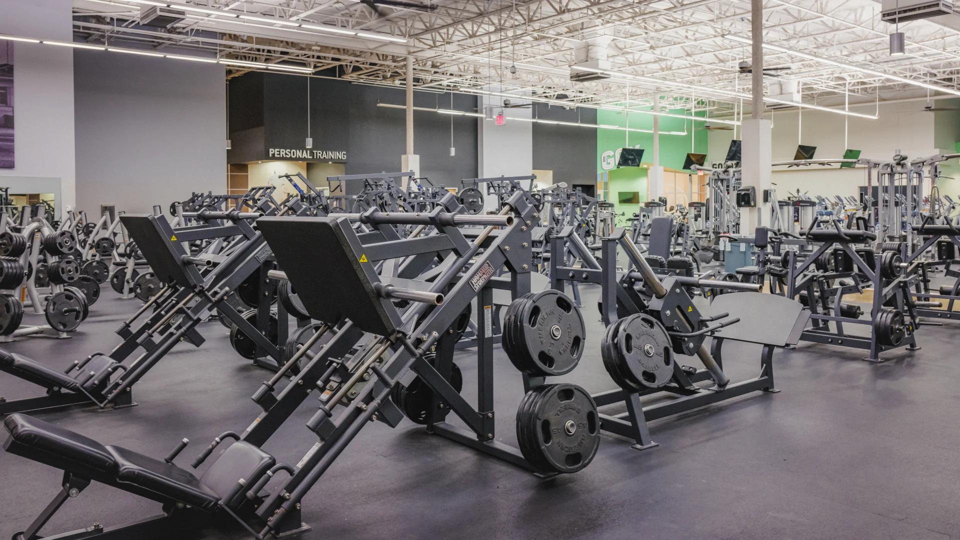 Free weights area at Gold's Gym Waxahachie featuring plate-loaded leg press machines and a variety of weightlifting equipment. The space has rubber flooring, bright overhead lighting creating a functional and spacious workout environment.