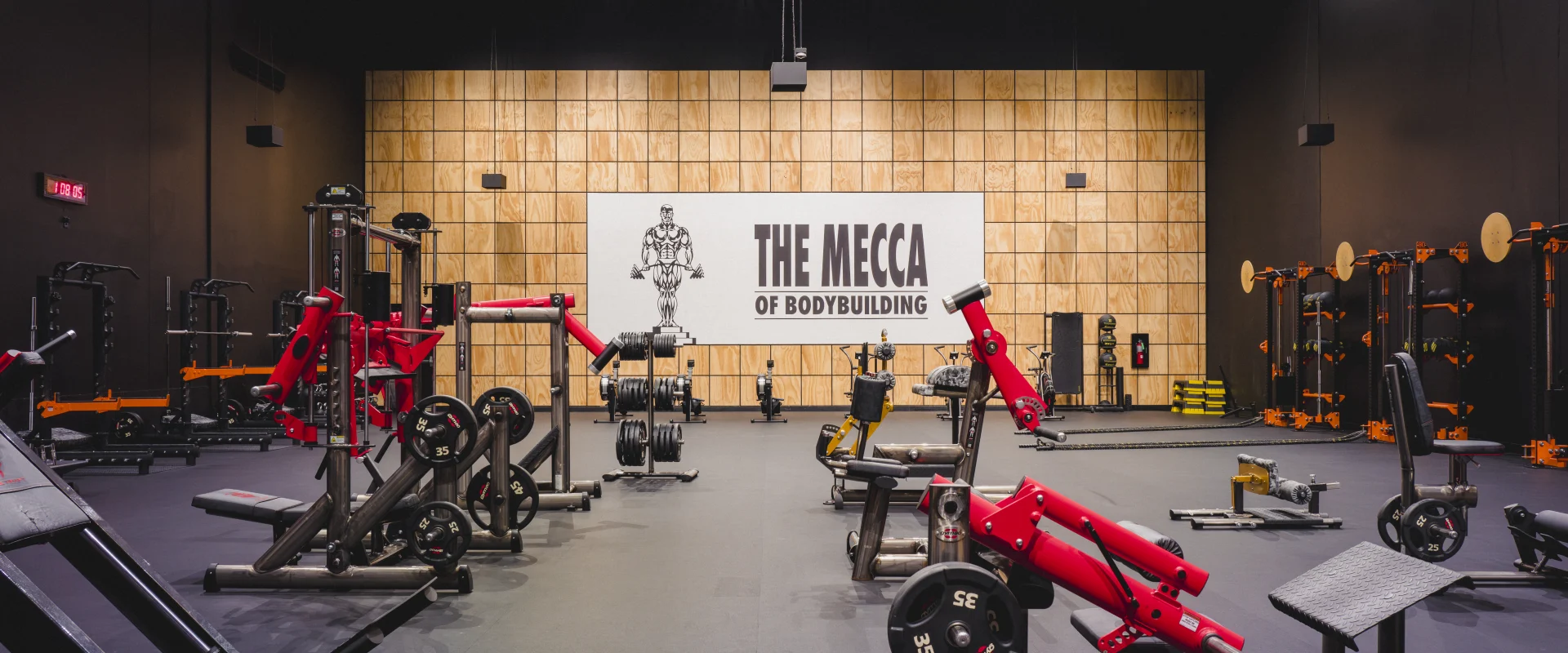 Wide-angle view of the power rack area at Gold’s Gym Little Elm featuring multiple power racks, weightlifting equipment, and a prominent "The Mecca of Bodybuilding" mural on a wooden accent wall. Bright overhead lighting highlights the black flooring and equipment layout.