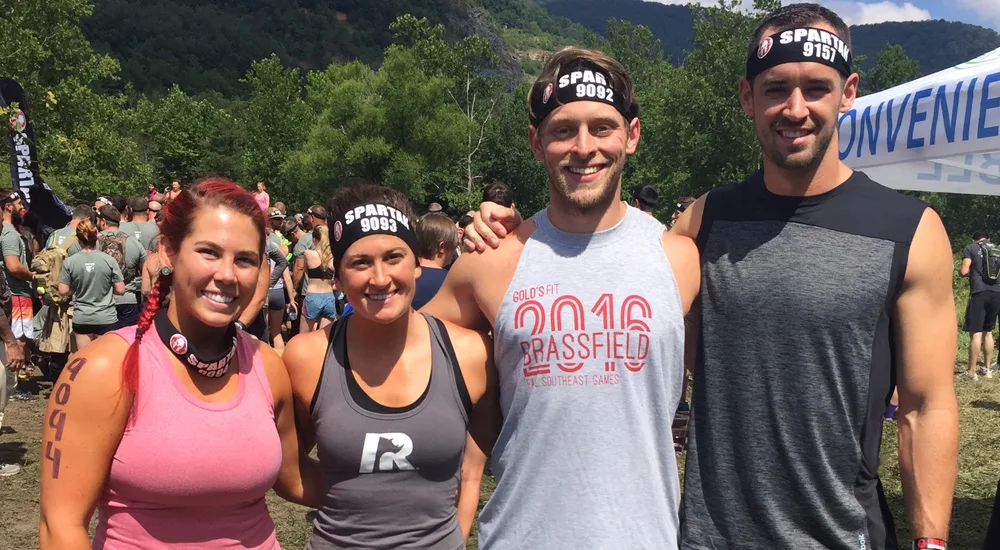 The image shows a group of people wearing matching sports jerseys, standing in a scenic outdoor setting with mountains and trees in the background.