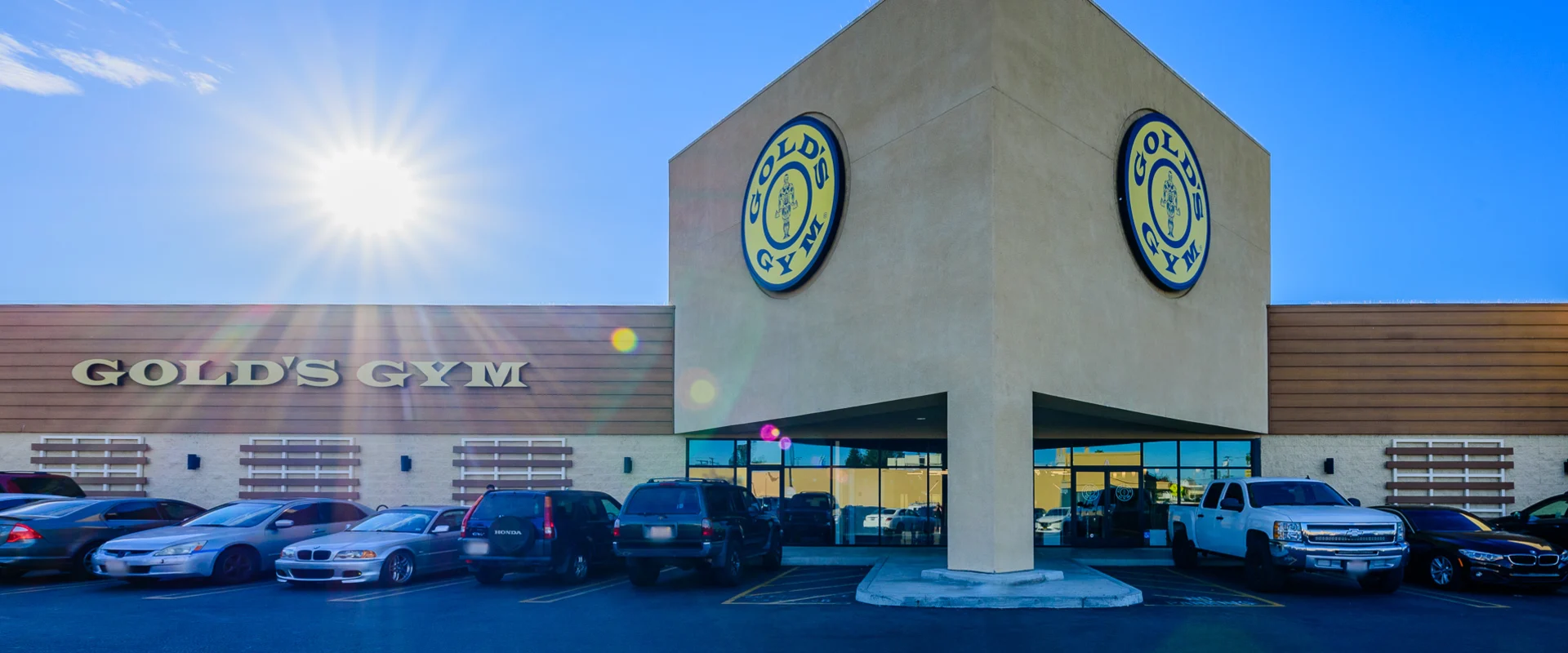 The exterior of Gold’s Gym Garden Grove showcases a beige stucco facade with the "Gold’s Gym" logo prominently displayed on two sections of the building. The sun shines brightly over the parking lot, casting lens flares across the image.
