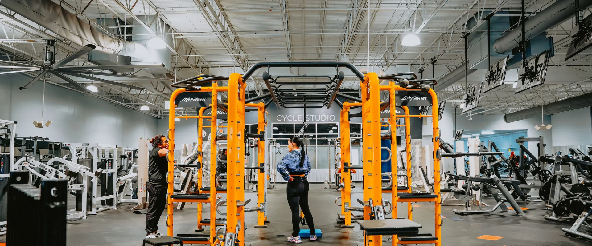 Functional training area at Gold’s Gym Tennessee, Hixson, featuring a vibrant yellow functional training rig equipped with pull-up bars and attachments, surrounded by benches and rubber flooring. The space includes a high ceiling with exposed beams and ducts, industrial-style lighting, and multiple overhead screens. A Cycle Studio entrance and various weight machines are visible in the background.