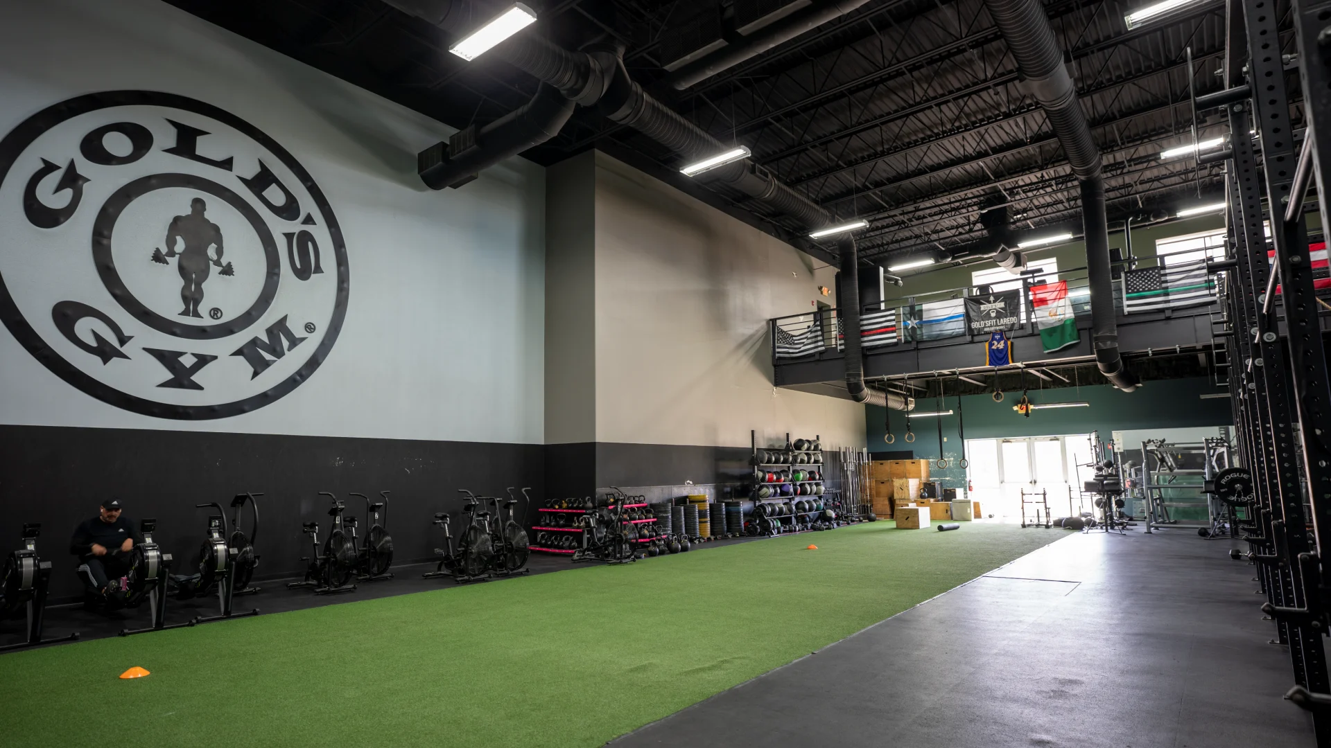 A large, open functional training area inside a Gold's Gym. The left wall features a massive black-and-white circular Gold's Gym logo above a row of rowing machines and stationary bikes. A long strip of green artificial turf runs down the center of the room, bordered by black rubber flooring and various equipment racks holding kettlebells, medicine balls, and weight plates. In the background, a mezzanine overlooks the floor, decorated with several flags.