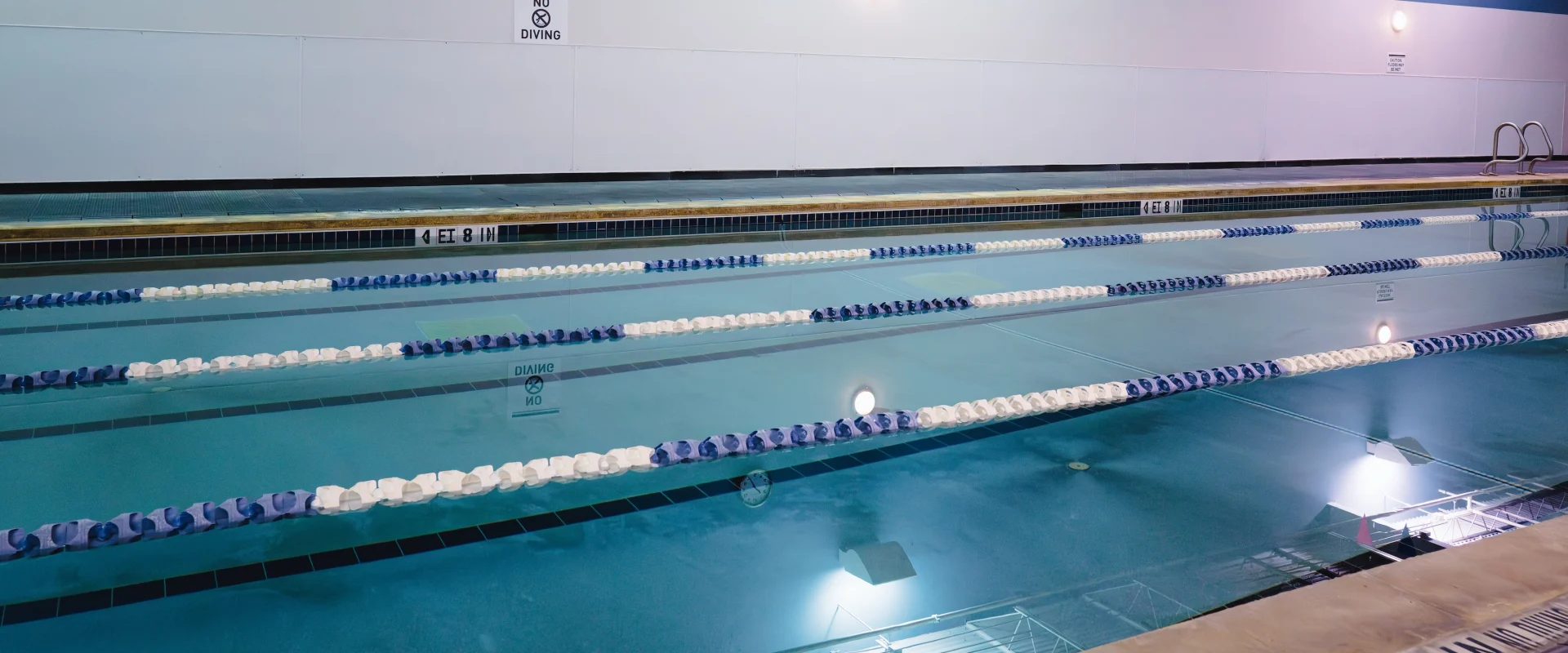 Indoor swimming pool with clear blue water, divided by lane markers. Blue and white walls with a "No Diving" sign create a serene atmosphere.